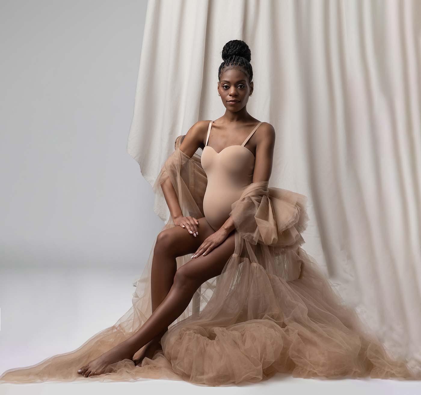 A professional photo of a pregnant woman wearing a long flowing fabric while sitting on a chair. The photo is tastefully lit by a maternity photographer.