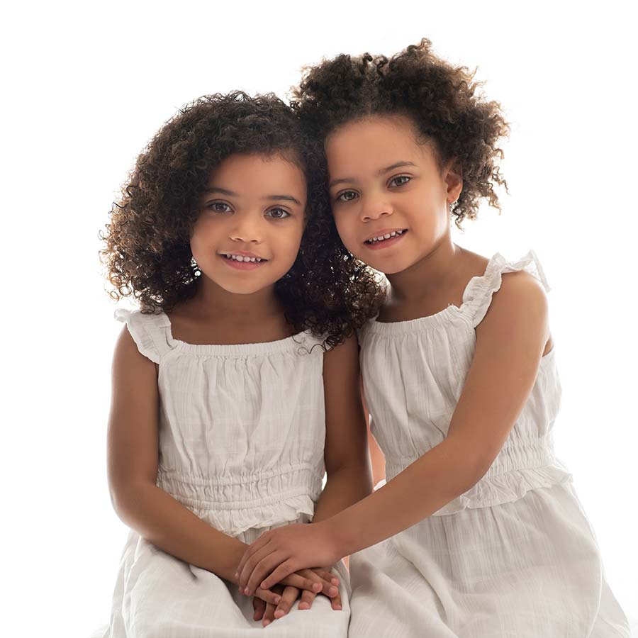 Two young sisters with curly hair and white dresses hold hands while posing at a baby photo studio in NYC. They're holding hands and leaning their heads together.