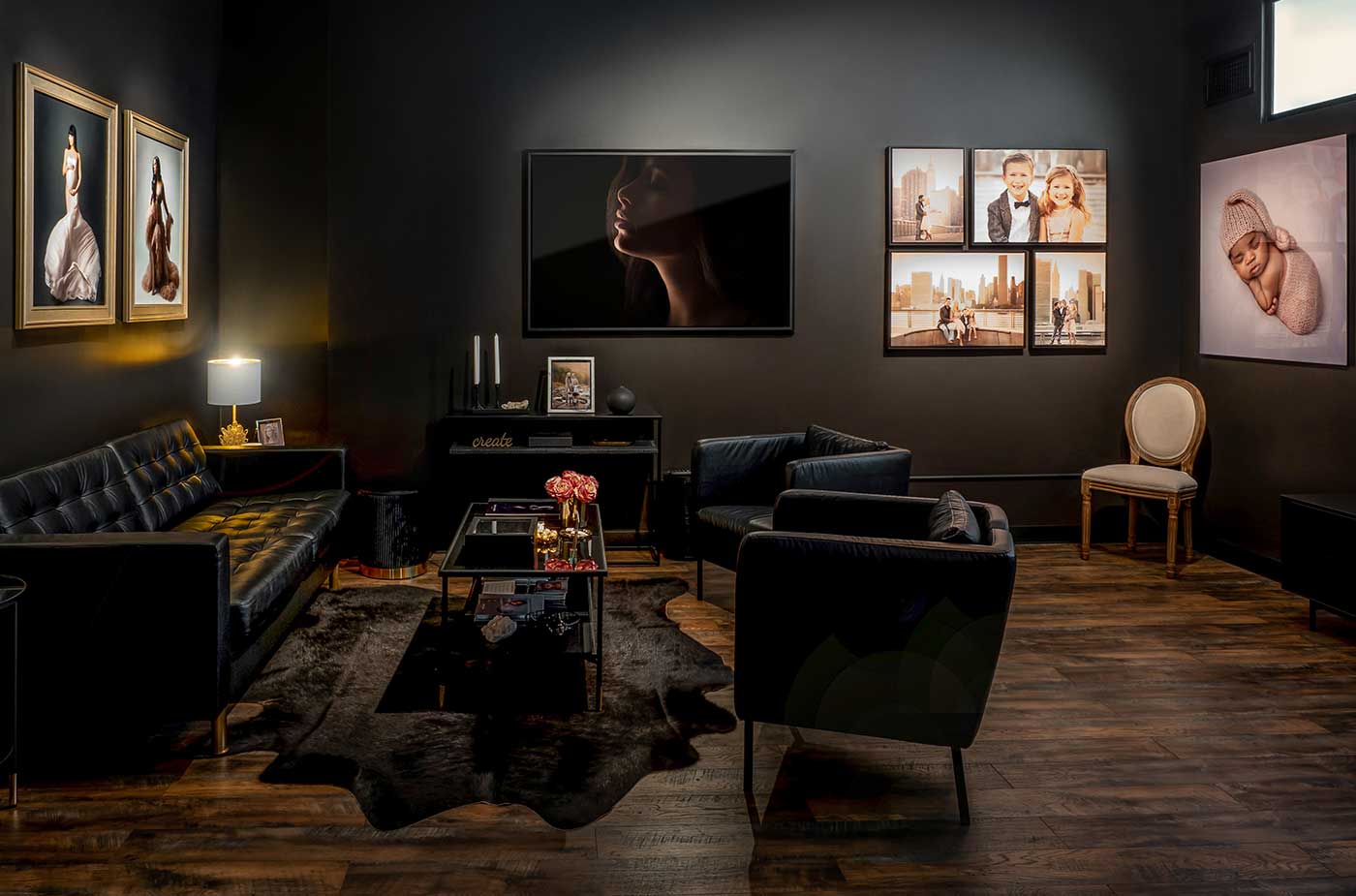 Reception area of a photography studio. A black leather couch with a glass coffee table dominates the scene. The walls are painted black and feature a variety of maternity, newborn, and baby photographs tastefully framed.