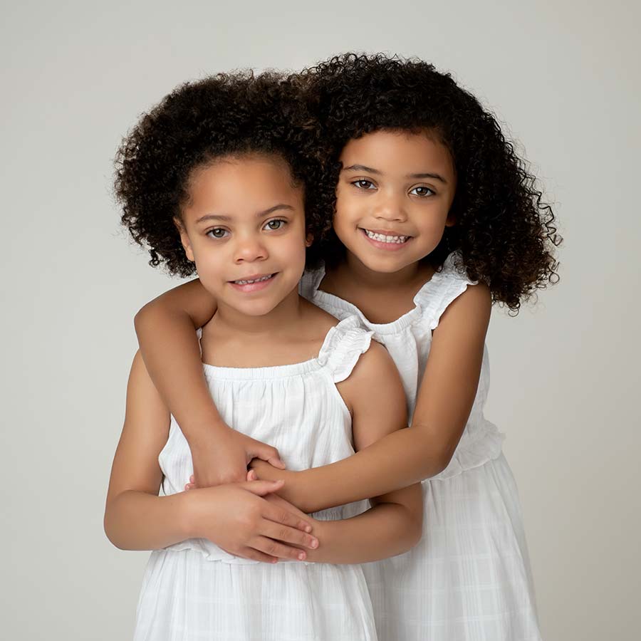 Two sisters, one standing behind the other with her arms wrapped around pose for a baby photographer. Their smiles are pleasant and convey happiness.