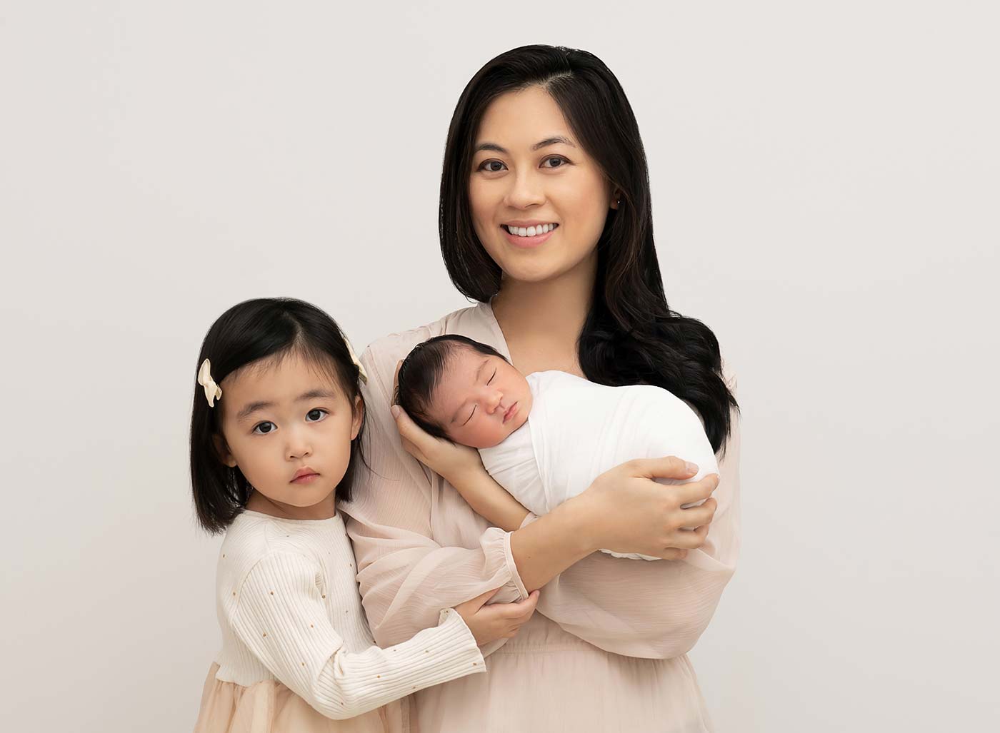 A mother, her daughter, and her infant baby pose for a family portrait at a newborn photo studio. They're dressed in neutral tones, with the baby swaddled in a white cotton cloth.