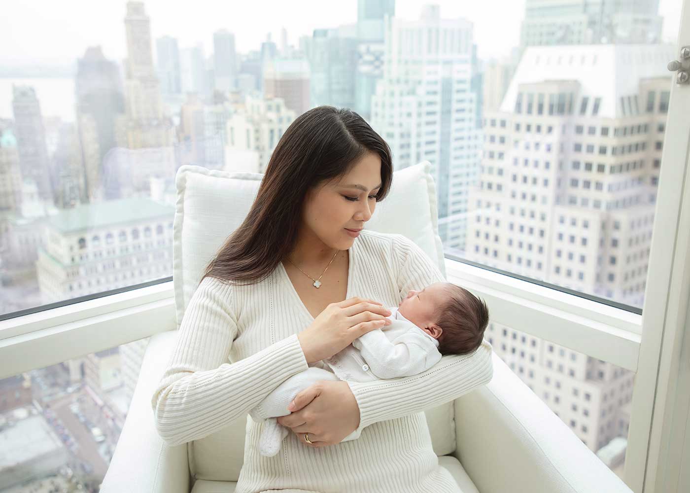 Mother holding her newborn baby on a couch. The background shows panoramic views of the NYC skyline, suggesting this image was taken at a high-rise apartment.