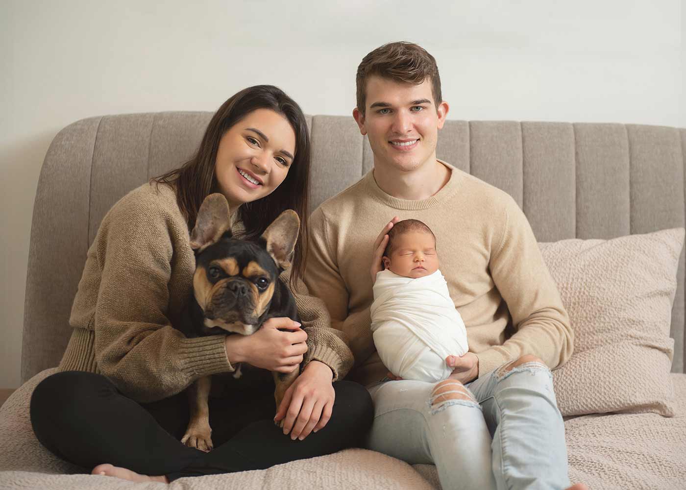 A family photo of a mother, father, their newborn baby, and a family pet. Both parents are dressed in casual wear, and their baby is swaddled in a white cotton cloth. They are all sitting on a couch in their NYC apartment.