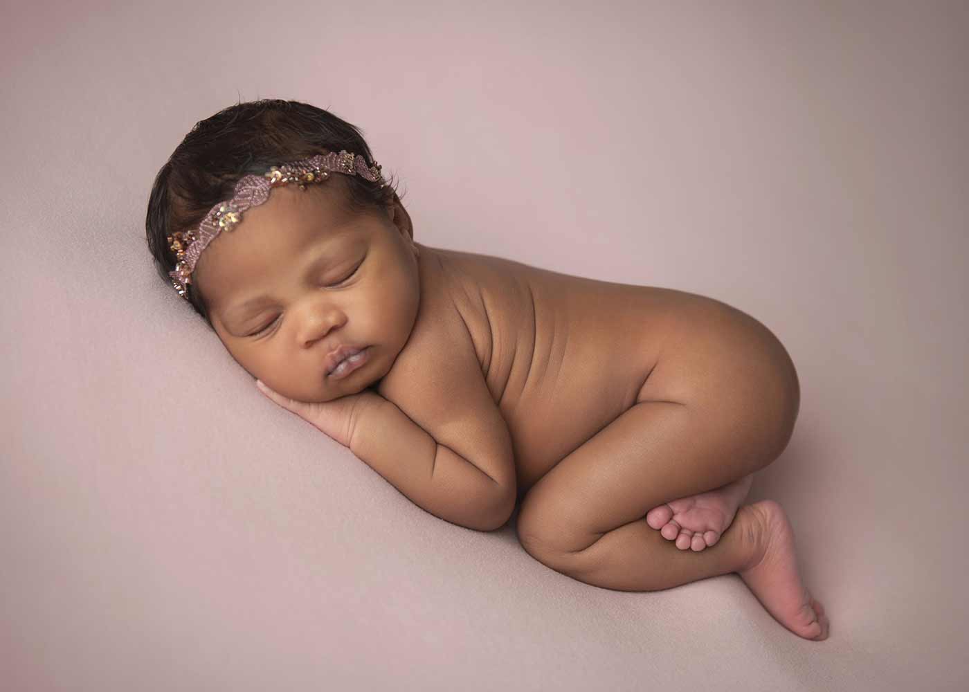 Newborn photo of a sleeping infant. The baby is wearing a decorative headband, and is beautifully posed on a pink blanket in a photo studio.