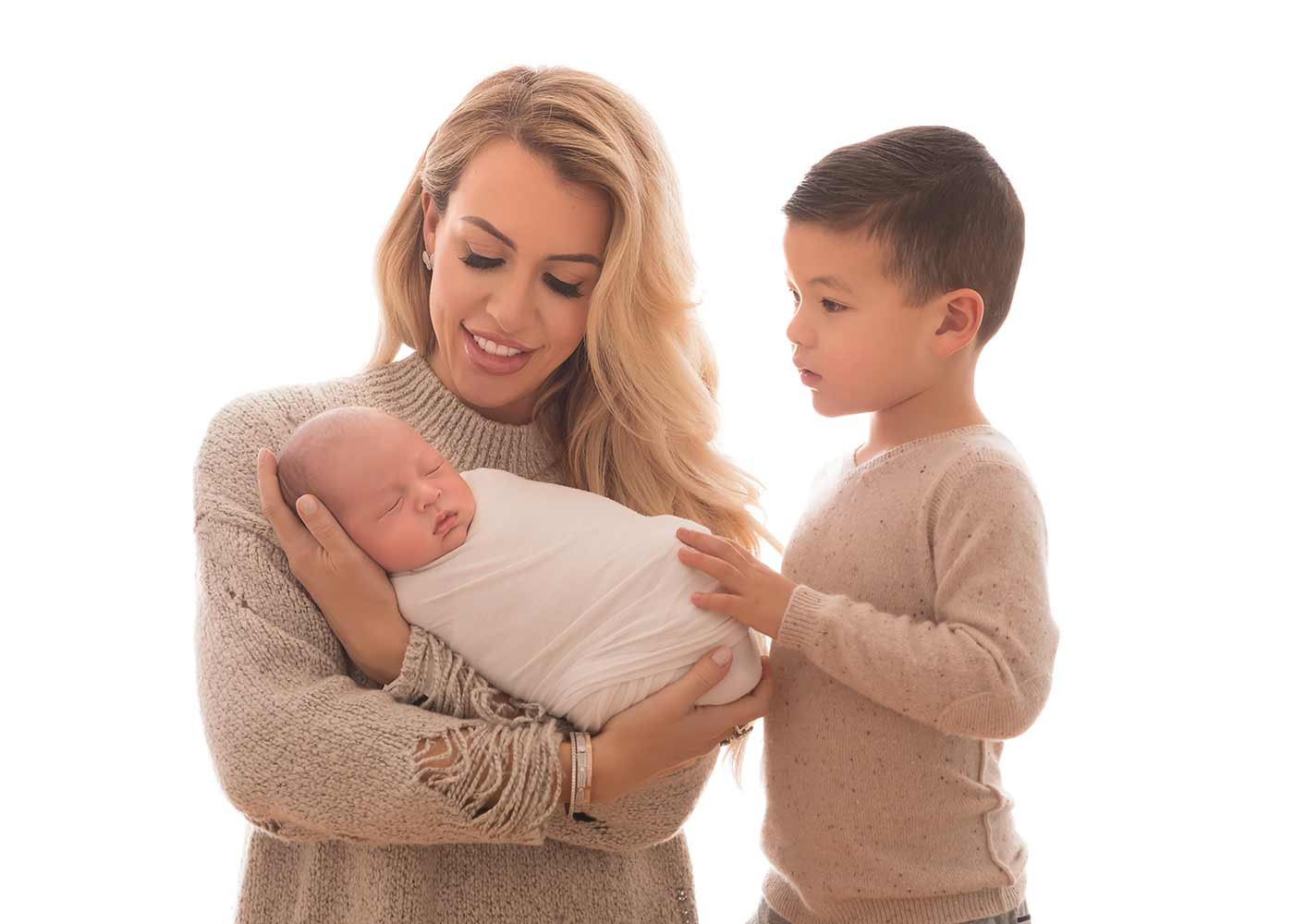 Mother holding her newborn baby. Her older son is gently petting his baby brother. The setting is reminiscent of a professional photo studio.