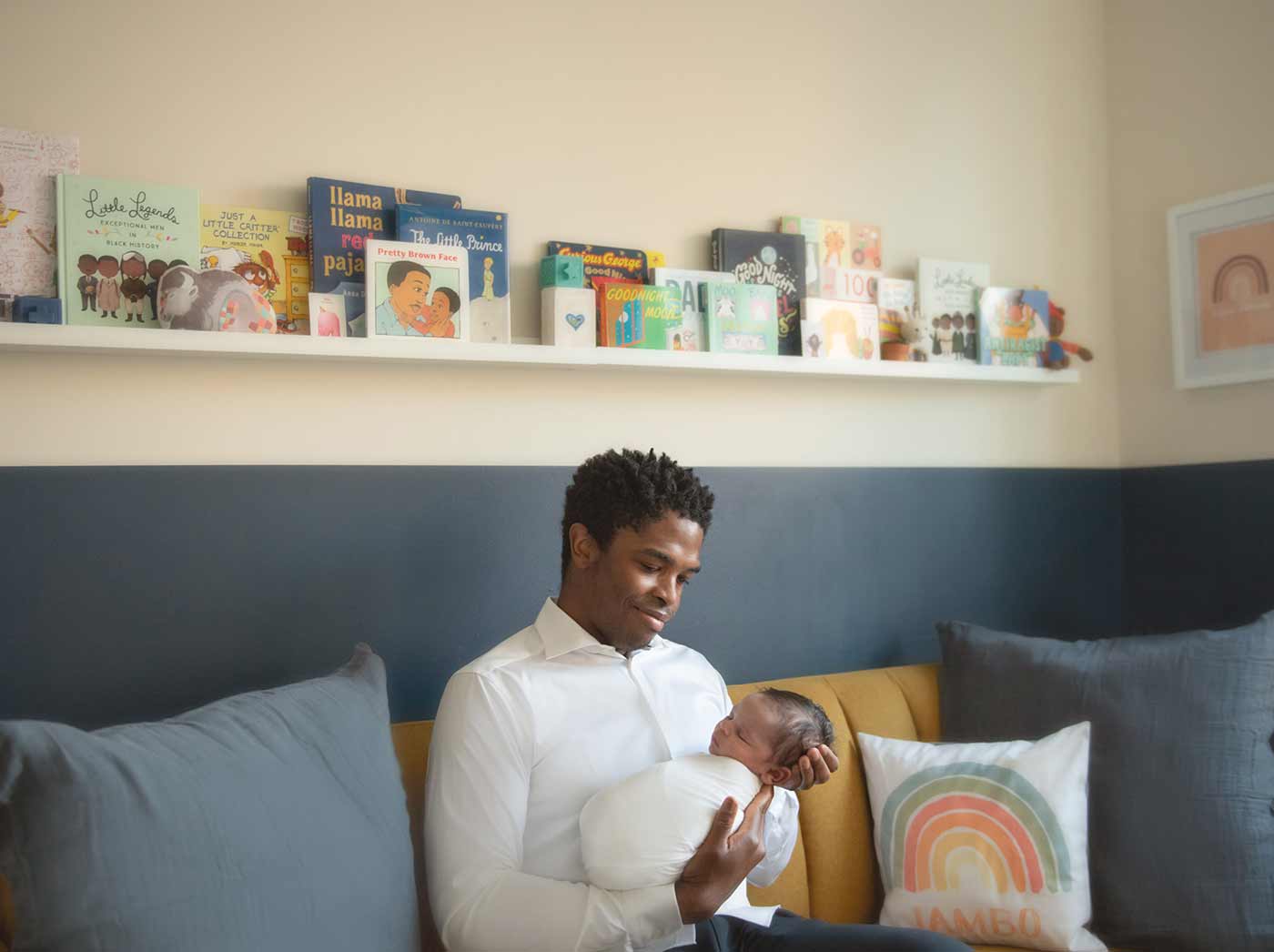 A lifestyle photo of a father holding his infant son in a nursery of their NYC apartment. The nursery is tastefully decorated with children's books and colorful pillows.