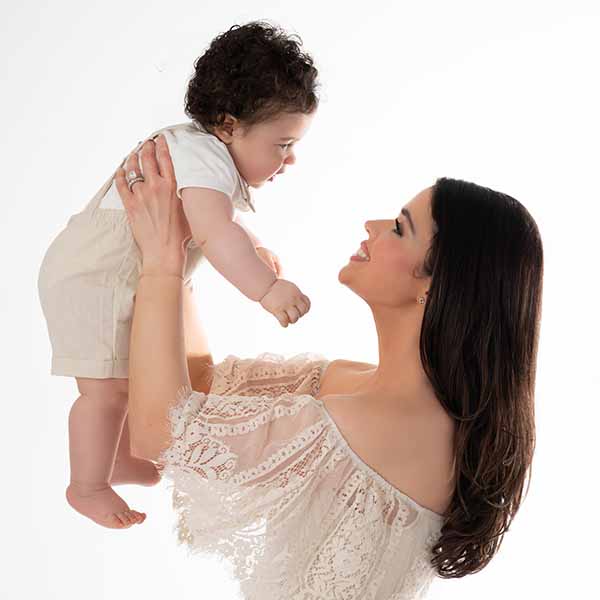 Mother holding up her baby boy. The two are looking at each other while wearing casual, off-white clothing. Their smiles indicate a happy moment.